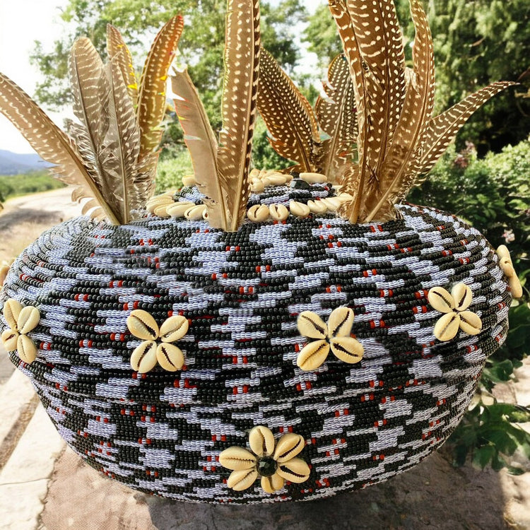 A beaded clay pot with black, white blue-striped beads and red accents. The pot has cowrie shells and feathers adorned on it, set outdoors against a natural backdrop.