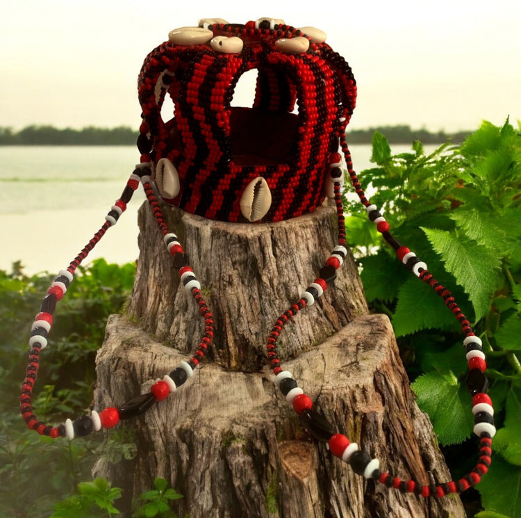 A handcrafted small beaded crown with red and black beads, displayed on a natural wooden stump with a body of water and greenery in the background.