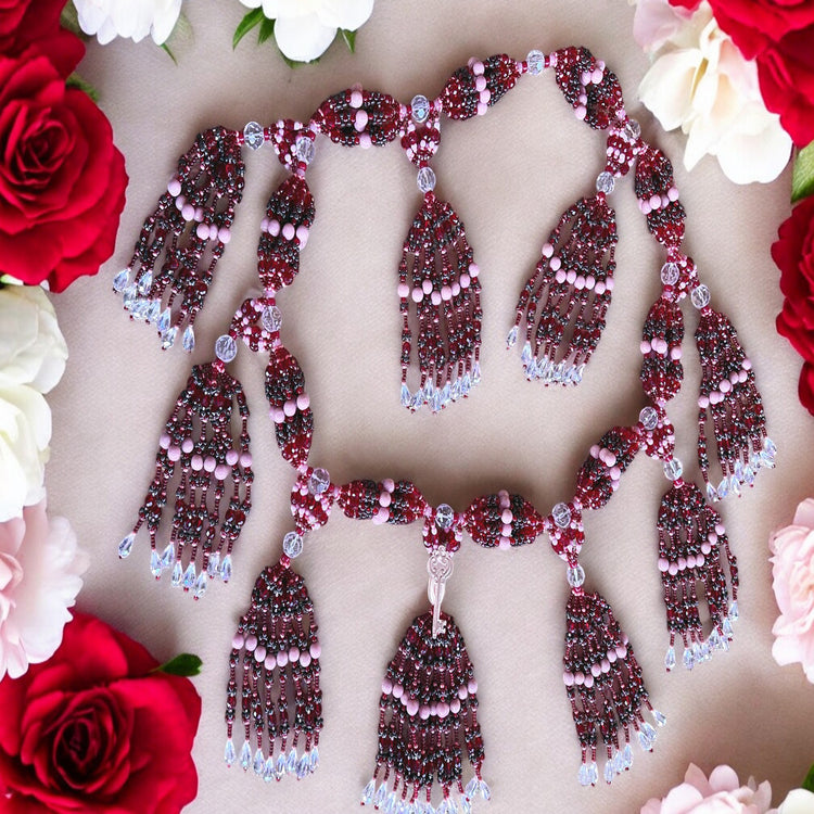 A beaded mazo (necklace) with earthy brown, soft pink, and deep red colors, adorned with a decorative key, displayed against a beige background surrounded by red and white flowers.