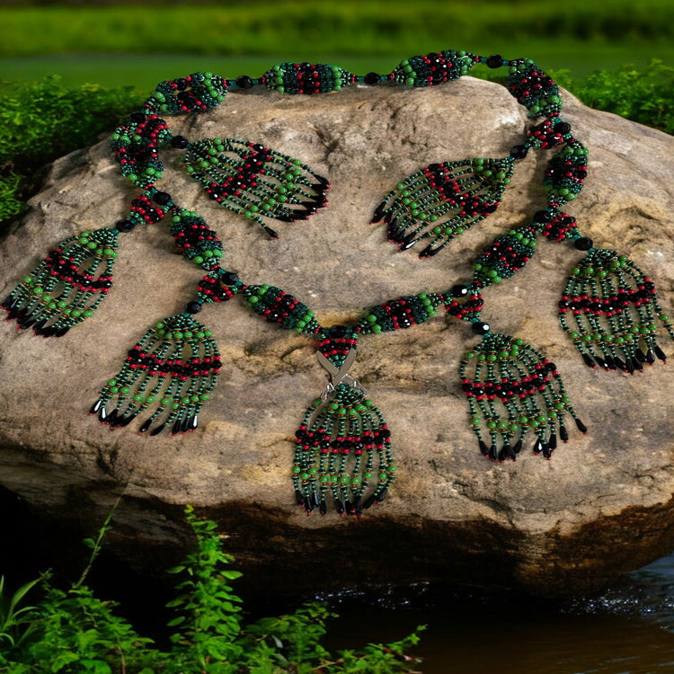 A beaded necklace with green, red and black beaded strings, and decorative accents shaped like crossed machetes, displayed on a rock.