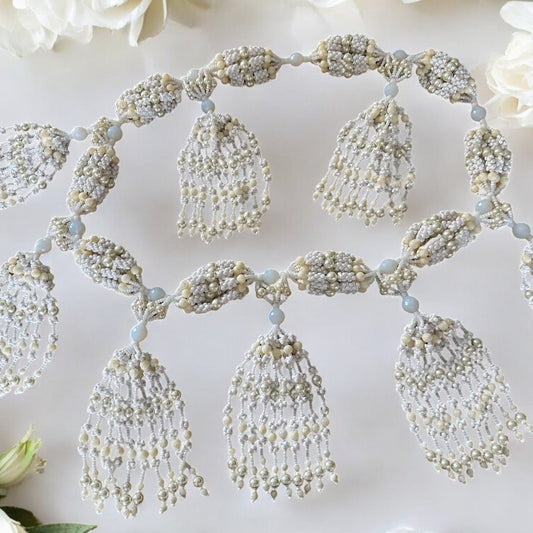 A detailed image of a Yoruba Oshanla-inspired mazo, featuring intricate beadwork with white, pearl, and mother of pearl accents, with a floral arrangement in the background.
