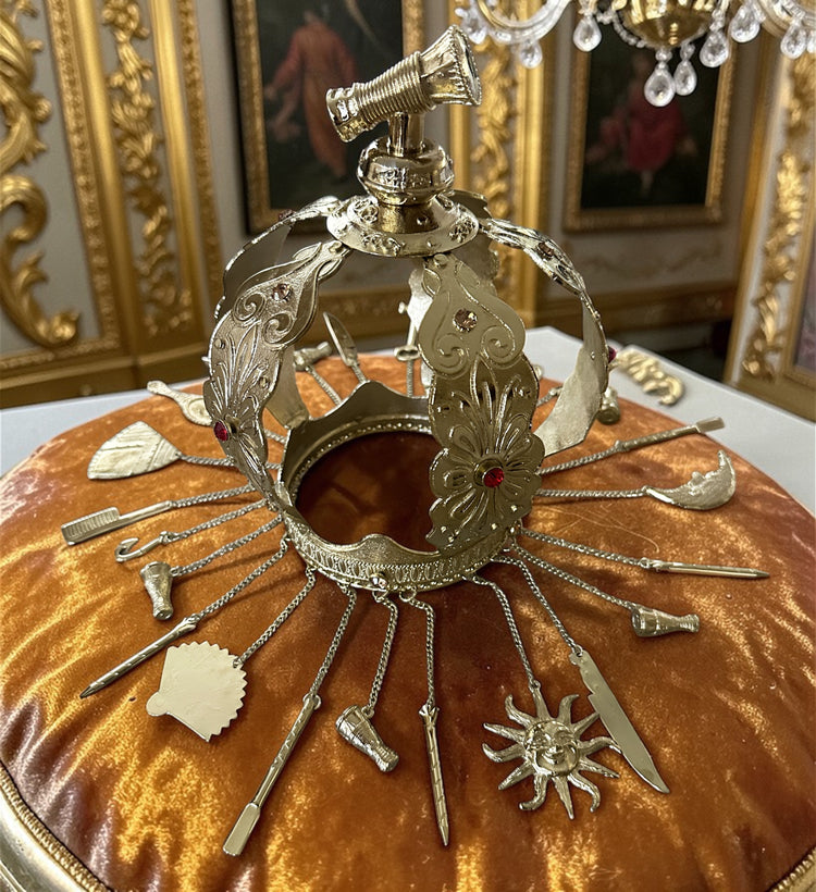 Decorative gold metal crown with adornments on a amber velvet pillow with ornate background.
