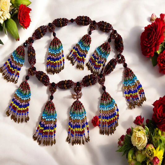 A colorful beaded necklace with a copper masquerade mask, displayed against a white background surrounded by red and yellow flowers.