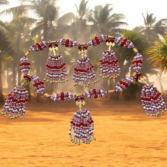 A Yoruba style glass mazo featuring red and white beads with gold accents, and six dual-head axe charms, displayed in an outdoor setting with palm trees in the background.
