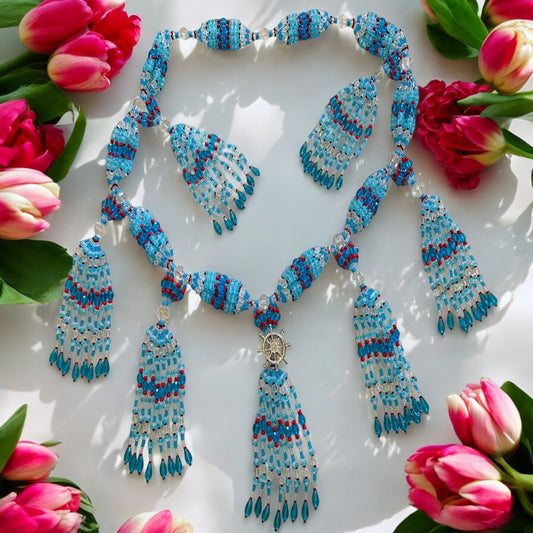 A detailed image of a glass beaded mazo, featuring a combination of aqua blue, light blue, red, transparent, and pink coral, arranged in a reflective surface with a symbolic boat wheel at the center, surrounded by pink coral.
