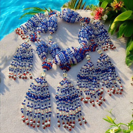 A beaded necklace set with navy blue, white, and red acrylic beads, displayed on a sandy surface with water in the background.