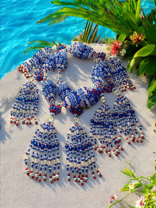 A beaded necklace set laid out on a sandy surface with a representation of Yemayá, featuring blue, red, and clear beads with silver accents, along with a body of water and greenery in the background.