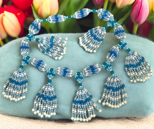 A beaded necklace with white, light blue, and aquamarine strings arranged in a symmetrical pattern with tassel-like ends, displayed against a floral background.