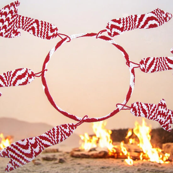 A beaded hoop of Shango's tools with red and white beads arranged in a pattern around a central point, with a blurred background of flames.
