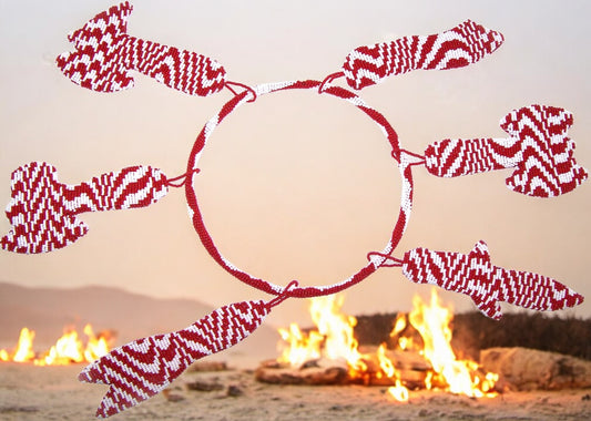 A beaded hoop of Shango's tools with red and white beads arranged in a pattern around a central point, with a blurred background of flames.