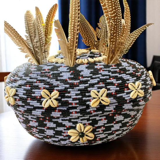 A beaded clay pot with black, white blue-striped beads and red accents, decorated with cowrie shells and feathers protruding from the top.