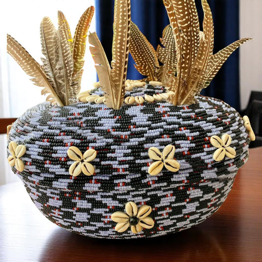 A beaded clay pot with black, white blue-striped beads and red accents, decorated with cowrie shells and feathers protruding from the top.