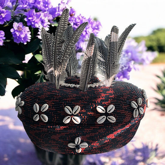 A beaded clay pot with brown and black colors, decorated with cowrie shells and feathers, displayed against a floral background.