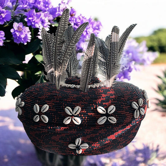 A beaded clay pot with brown and black colors, decorated with cowrie shells and feathers, displayed against a floral background.