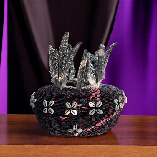 A beaded clay pot with a geometric pattern using brown and black beads, and cowrie shells, displayed on a wooden surface with a purple background.