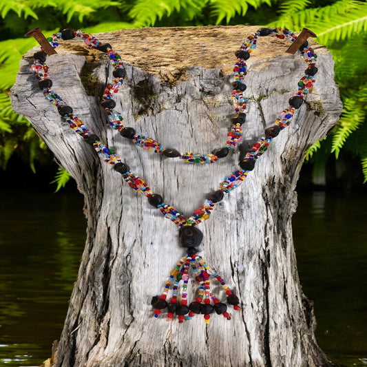 A multi-colored 4-strand necklace with black skull accents displayed on a wooden stump with a body of water and greenery in the background.