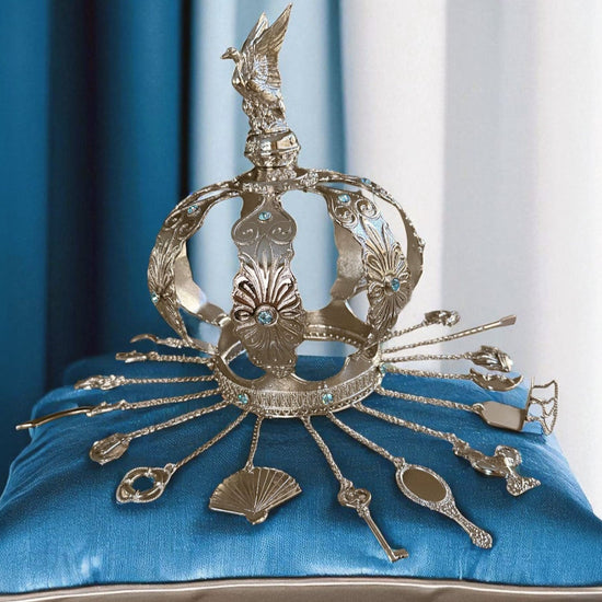 Decorative silver crown with intricate designs on a blue cushion against a blue curtain background