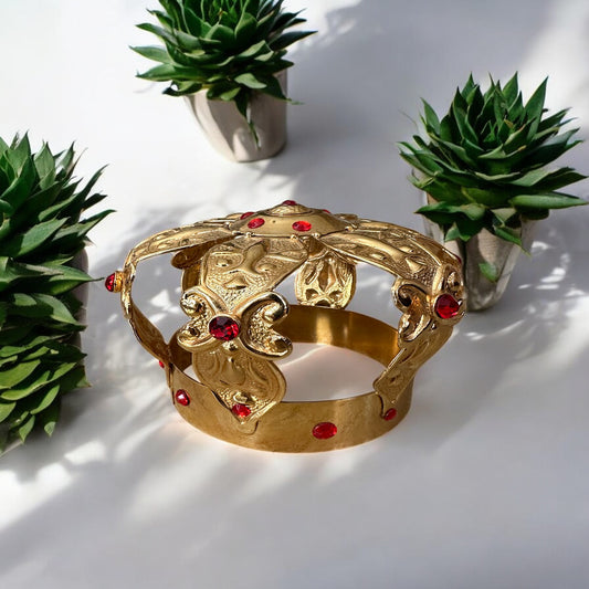 A gold-colored metal crown with ruby accents, displayed on a white surface with green plants in the background.