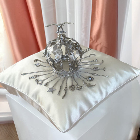 Decorative silver colored crown on a white cushion with pink curtains in the background