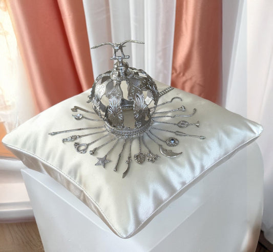 Decorative silver colored crown on a white cushion with pink curtains in the background