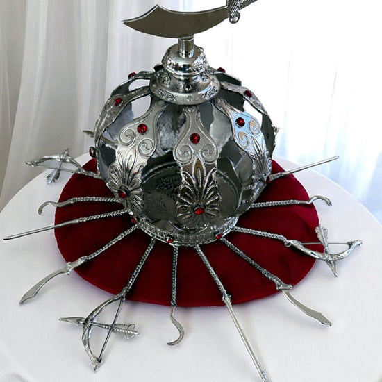Decorative silver metal crown with red gemstones and a sabre topper on a white background