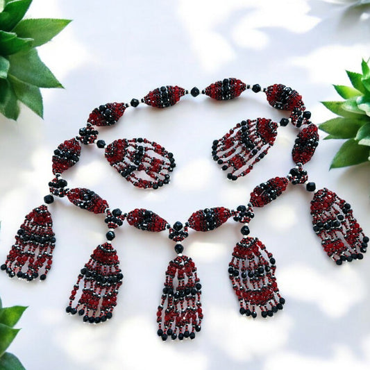 A decorative acrylic necklace for Elegua featuring red, black, and white beads with tassel details, displayed against a white background with green leaves.