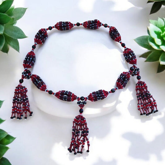 A beaded mazo necklace featuring red and black glass beads, with intricate designs and tassel details, displayed against a white background with green foliage.