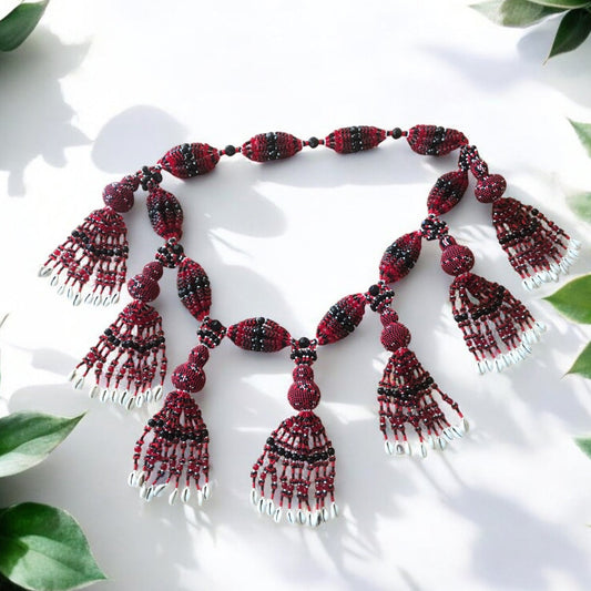 A handcrafted 10-string Eleguá mazo necklace featuring red, black, and white beads, and small beaded gourds, displayed on a white background with green leaves.