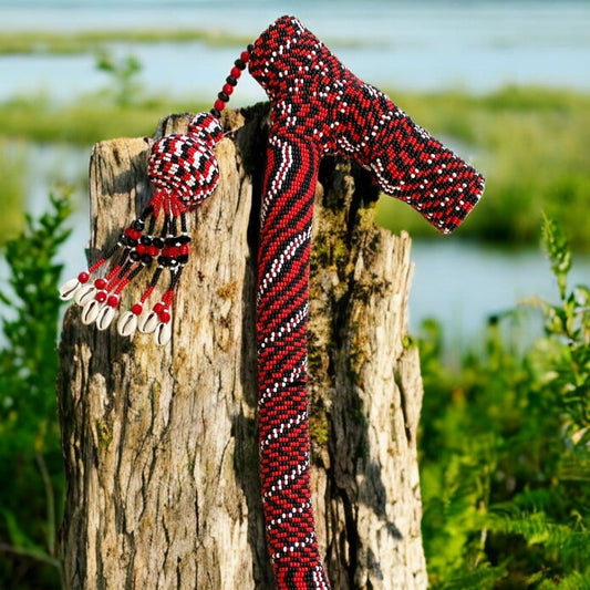 A handcrafted beaded grapnel made from the limb of a guava tree, featuring red and black beads and a small beaded gourd, displayed on a wooden post with a natural background.