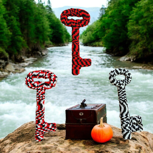 Three beaded skeleton keys in red and black patterns, displayed on a rock beside a small wooden box and a pumpkin, with a river in the background.