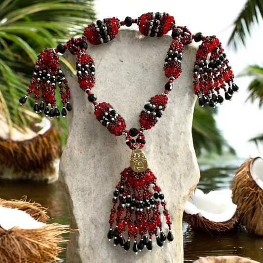 A beaded necklace with red, black, and white strings, displayed on an upright stone, set against a tropical backdrop with coconuts and greenery.