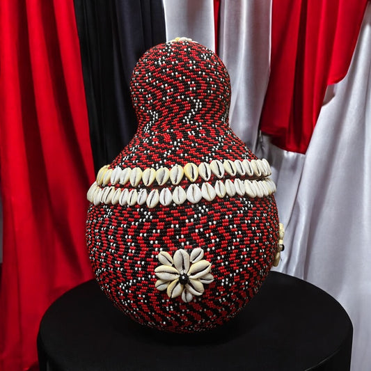 A large, beaded gourd with a red and black pattern, decorated with white beads and a cowrie floral design, displayed on a stand.
