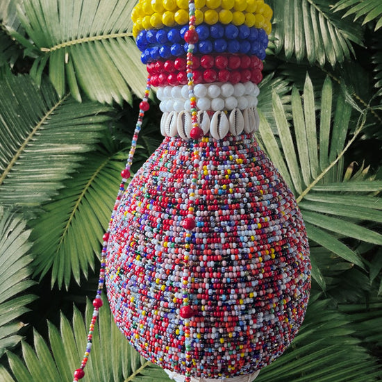 A handcrafted, multicolored beaded gourd with a design featuring yellow, blue, red, and white beads on the neck, displayed against a foliage background.