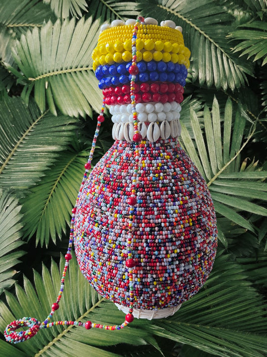 A handcrafted, multicolored beaded gourd with a design featuring yellow, blue, red, and white beads on the neck, displayed against a foliage background.