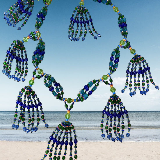 A beaded mazo necklace featuring multiple strands with blue and green beads, displayed against a beach backdrop.