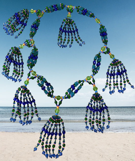 A beaded mazo necklace featuring multiple strands with blue and green beads, displayed against a beach backdrop.