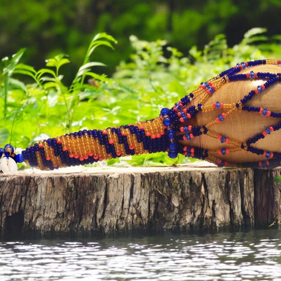 A beaded maraca with a wooden handle, featuring blue, topaz, and coral beads, resting on a wooden stump with water in the background.