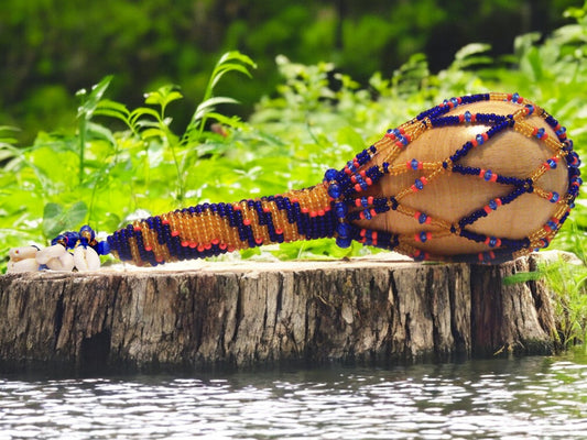 A beaded maraca with a wooden handle, featuring blue, topaz, and coral beads, resting on a wooden stump with water in the background.
