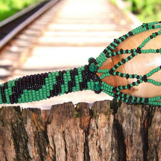 A beaded maraca with green and black colors, adorned with Czech glass beads, displayed on a wooden surface with a train track in the background.