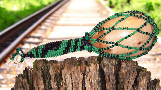A beaded maraca with green and black colors, adorned with Czech glass beads, displayed on a wooden surface with a train track in the background.