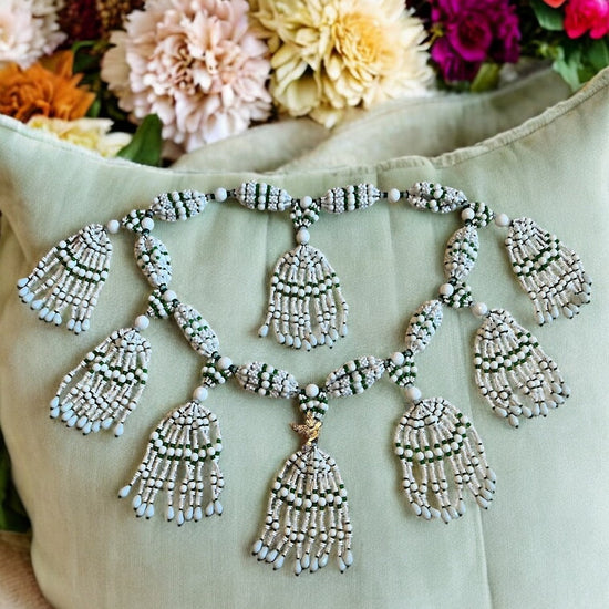 A detailed image of a Yoruba-style mazo, featuring multiple strands of white and green beads with decorative dove accents, displayed on a cushion with floral patterns in the background.