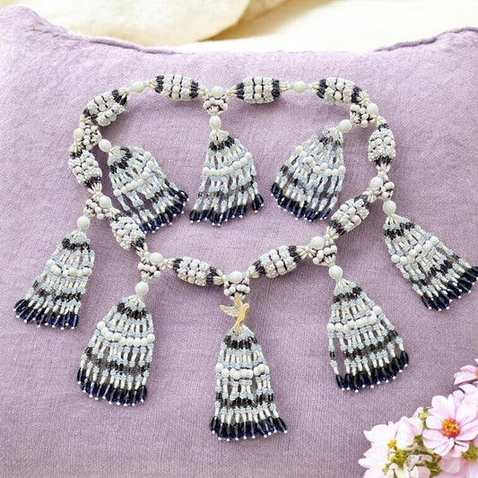 A handcrafted beaded necklace with white and purple strings, arranged in a decorative pattern, displayed on a purple cushion with a dove shaped pendant at the center.