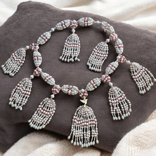 A detailed image of a 10-string mazo necklace with white and coral beads, featuring a decorative dove accent, displayed on a dark cushion.
