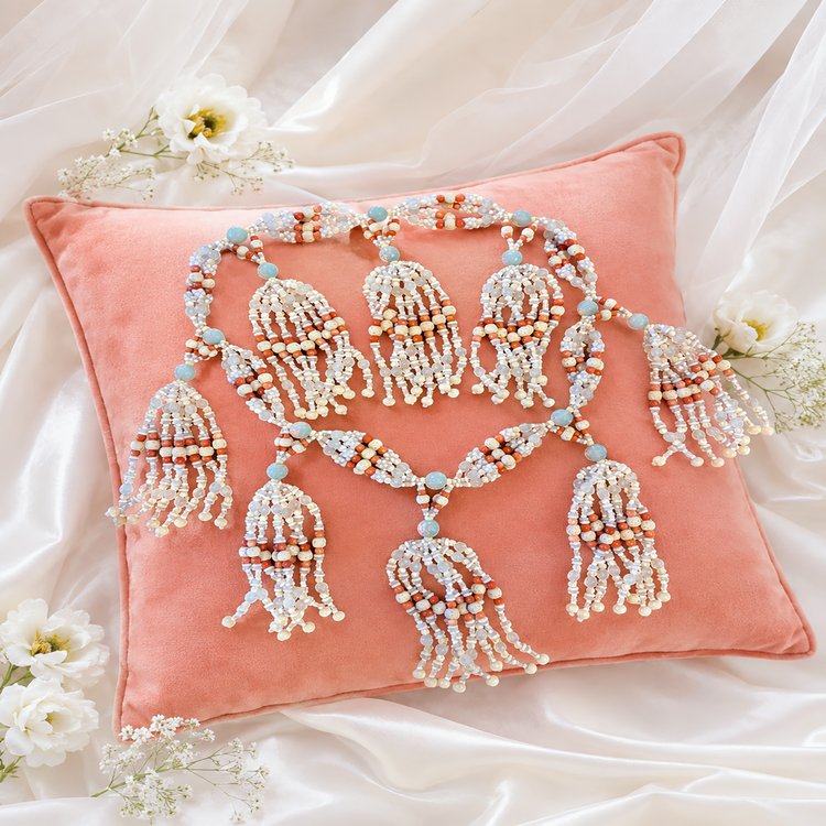 A coral colored velvet throw pillow decorated with a large, ornate beaded tassel garland in white, bone, opal and coral colored  beads, styled on draped white satin fabric with small white flowers.