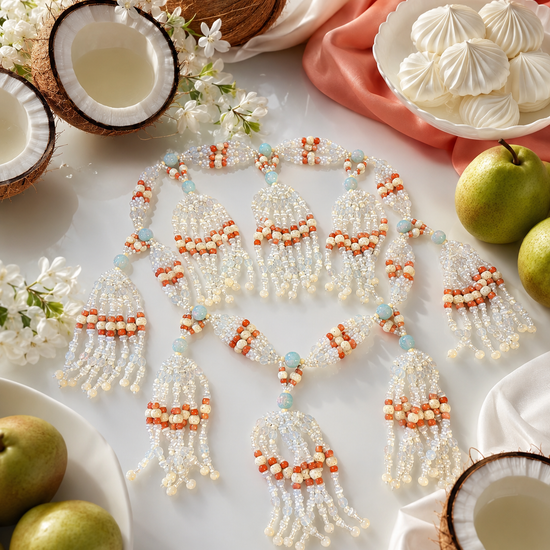 A beaded tassel necklace with white and coral, bone and opal beads. Styled on a white surface with coconuts, pears, white meringues, and white flowers.