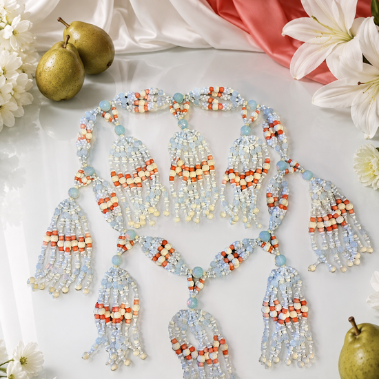 A beaded tassel necklace laid flat on a white surface, decorated with a large, ornate beaded tassel garland in white, bone, opal and coral colored  beads, styled on draped white satin fabric with small white flowers. The necklace is styled with white lilies, pears, and draped satin fabric in the background.