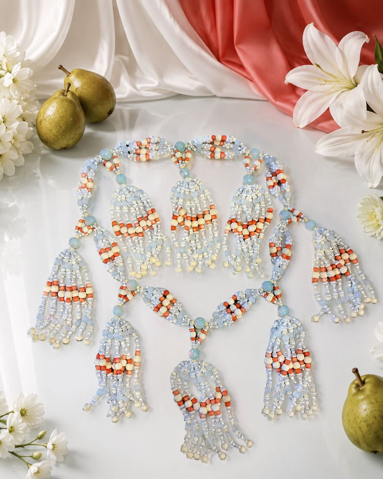 A beaded tassel necklace laid flat on a white surface, decorated with a large, ornate beaded tassel garland in white, bone, opal and coral colored  beads, styled on draped white satin fabric with small white flowers. The necklace is styled with white lilies, pears, and draped satin fabric in the background.