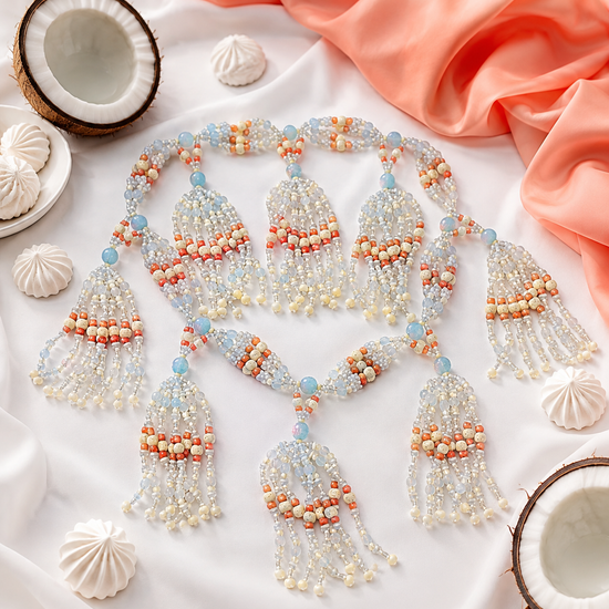 A beaded tassel necklace with intricate patterns of  white, bone, opal and coral colored  beads,  styled on a white fabric background with decorative seashells and coral fabric.
