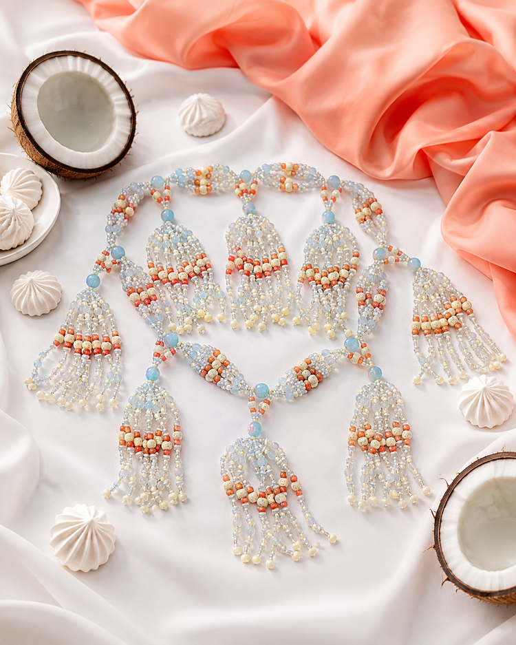 A beaded tassel necklace with intricate patterns of  white, bone, opal and coral colored  beads,  styled on a white fabric background with decorative seashells and coral fabric.