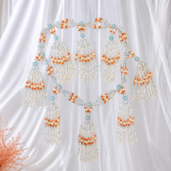 An  ornate beaded necklace with tassels  in white, bone, opal and coral colored  beads,  arranged against a white draped fabric background.
